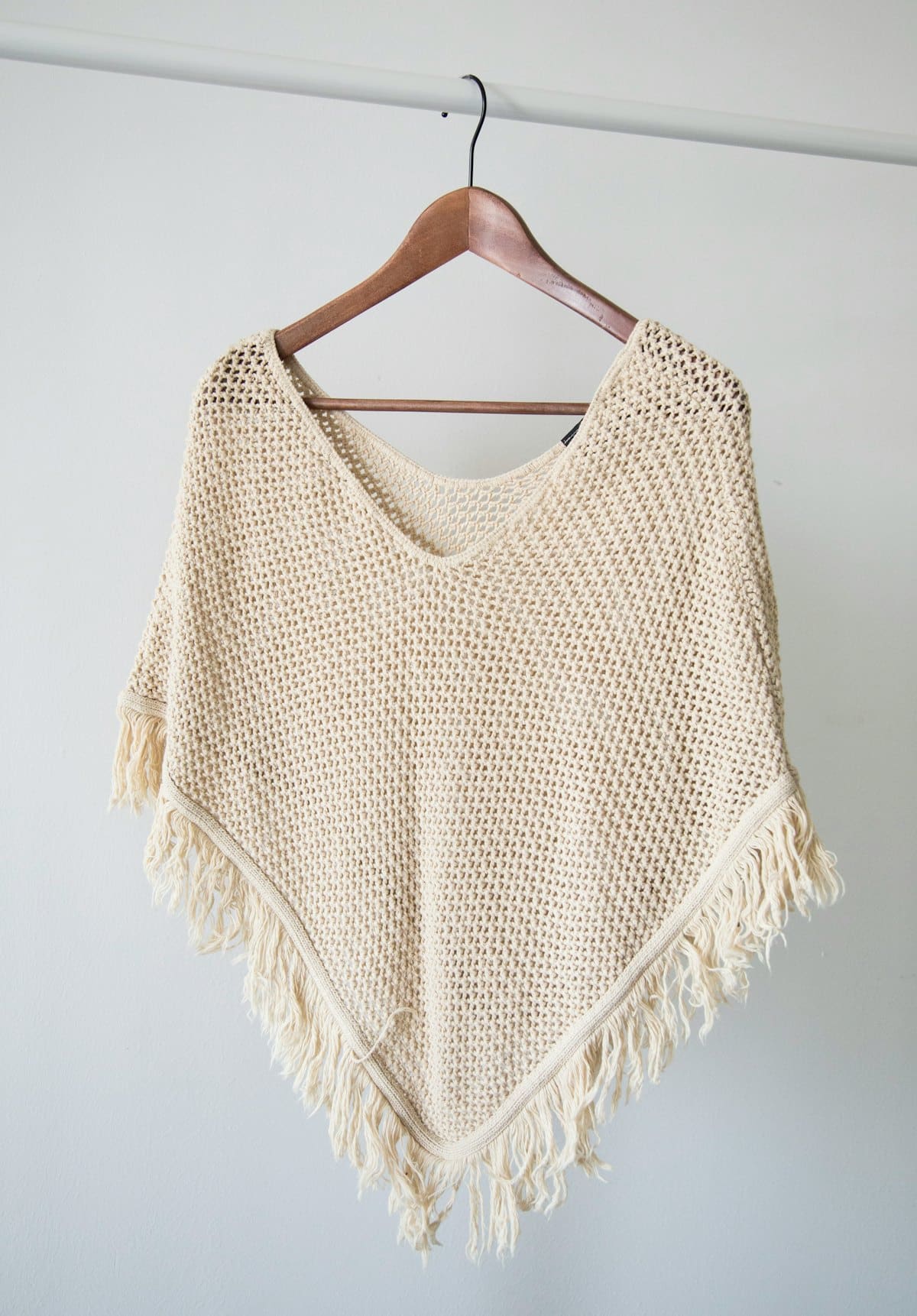 Hand-crocheted ecru poncho with fringe hanging on a wooden hanger